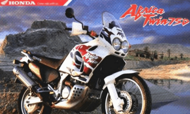 A Honda Africa Twin 750 motorcycle, symbolizing the evolution of adventure motorcycles in South Africa, is parked on sandy, rocky terrain against a backdrop of hills and blue sky. The bike showcases a striking white, red, and purple color scheme with visible branding.