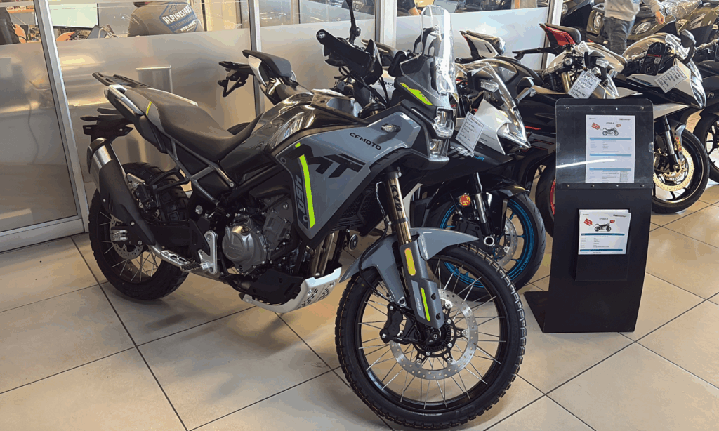 A black and green CFMOTO adventure bike is on display in a showroom in South Africa, positioned among other motorcycles with sales information attached and stands nearby. The well-lit setting features glass panels in the background.