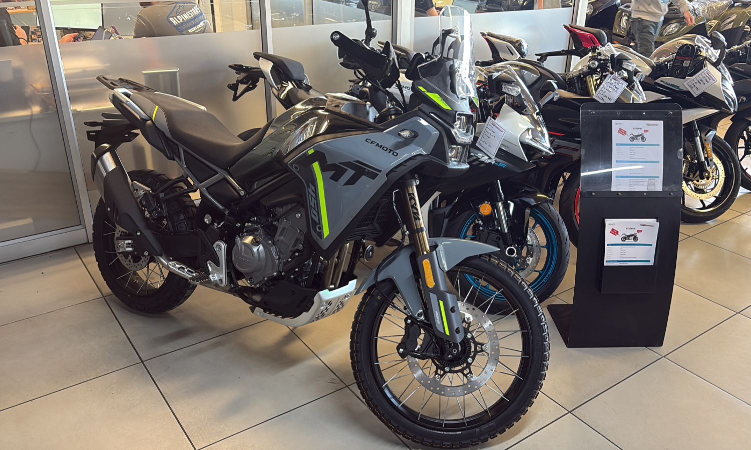 A black and green CFMOTO adventure bike is on display in a showroom in South Africa, positioned among other motorcycles with sales information attached and stands nearby. The well-lit setting features glass panels in the background.