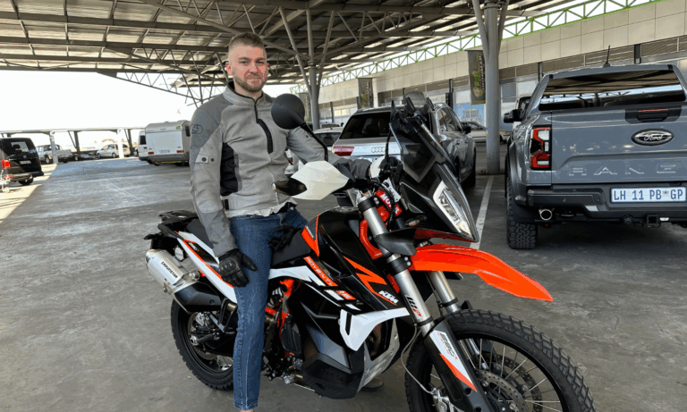 A man wearing a gray motorcycle jacket and jeans sits on an orange and black KTM adventure motorcycle—known for its reliability issues—in a covered parking area with cars and trucks in the background.