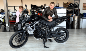 A man in a brown leather jacket leans on a black Triumph Tiger 800 XCx inside a showroom, while another person stands in the background looking at a phone. Tires and motorcycle gear are visible on shelves behind them.