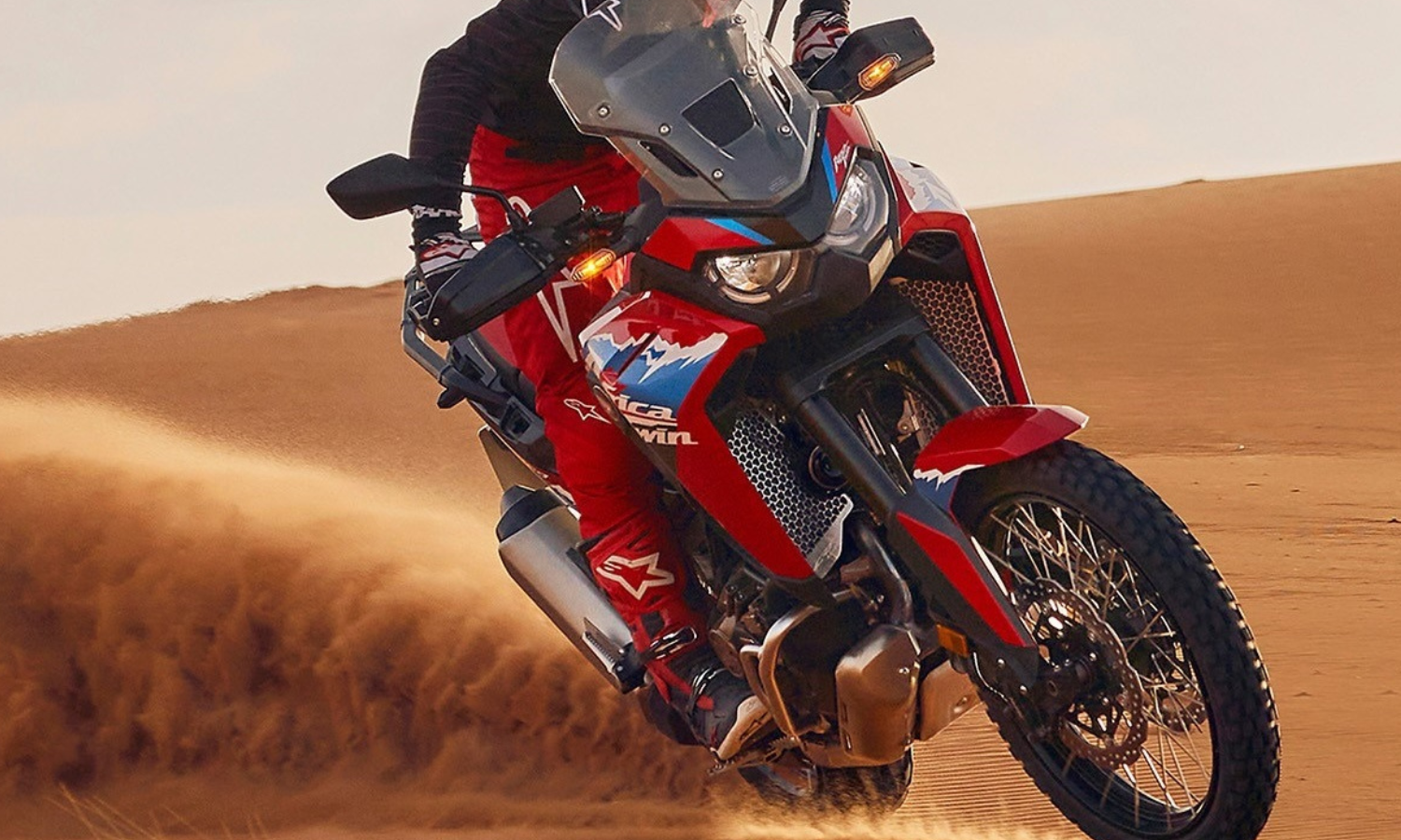 A motorcyclist in red gear rides a Honda Africa Twin CRF1100 adventure motorcycle through sandy desert terrain, kicking up dust behind the bike.