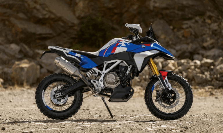 A BMW Concept F 450 GS adventure bike in blue, white, and red with off-road tires is parked on a rocky dirt surface, set against a blurred rocky background.