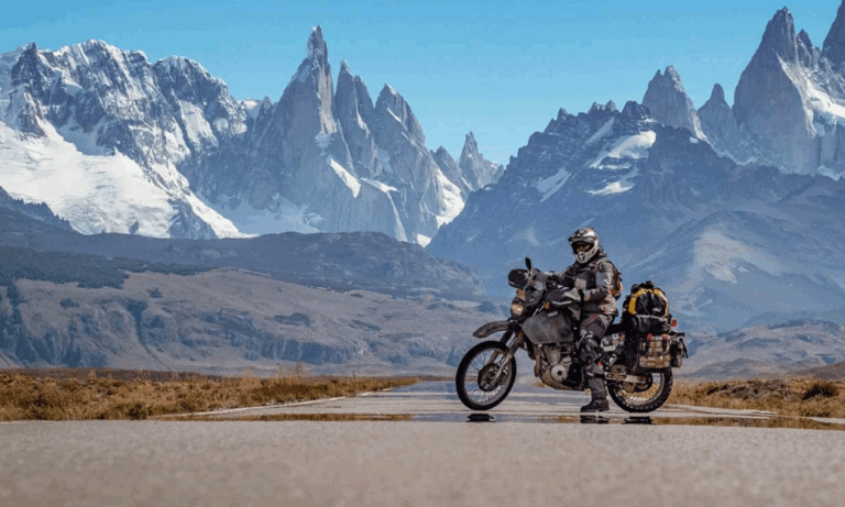 A motorcyclist in full gear sits on a loaded adventure bike on an empty road, ready for the Turkana Gear Challenge Safari 2024, with dramatic, jagged snow-capped mountains and clear blue sky in the background.