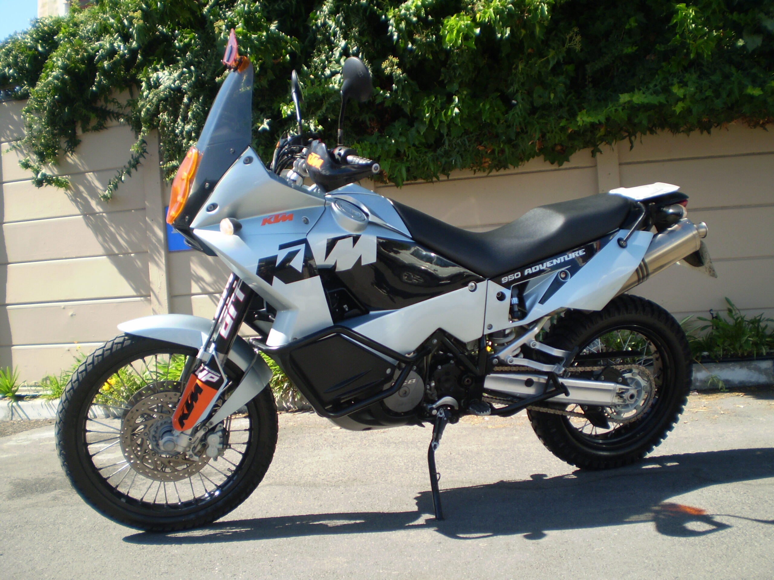 A side view of a KTM 950 Adventure motorcycle parked in front of a wall covered with greenery.