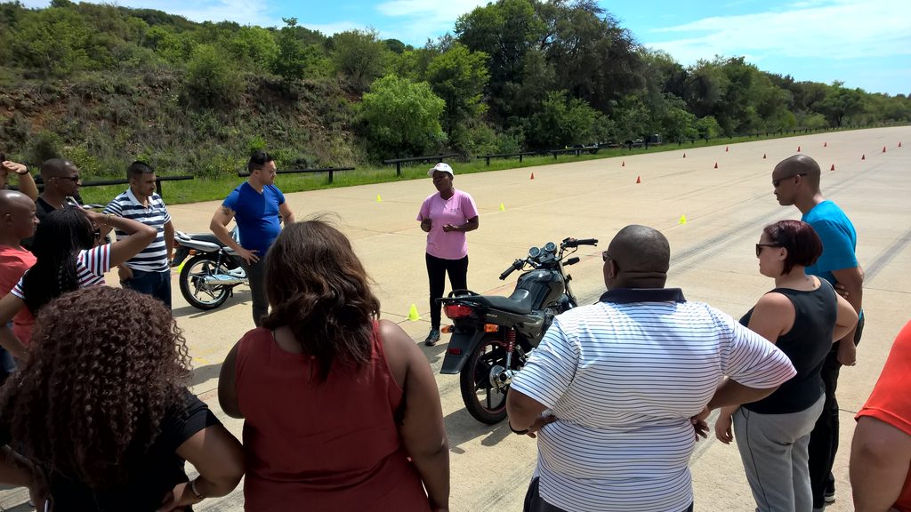 An instructor briefs a group of new riders beside a training motorcycle on the cones course