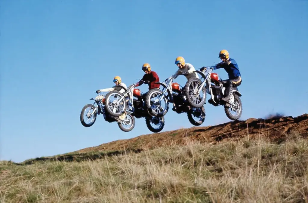 Four riders jumping vintage dirt bikes over a grassy hill in 1970