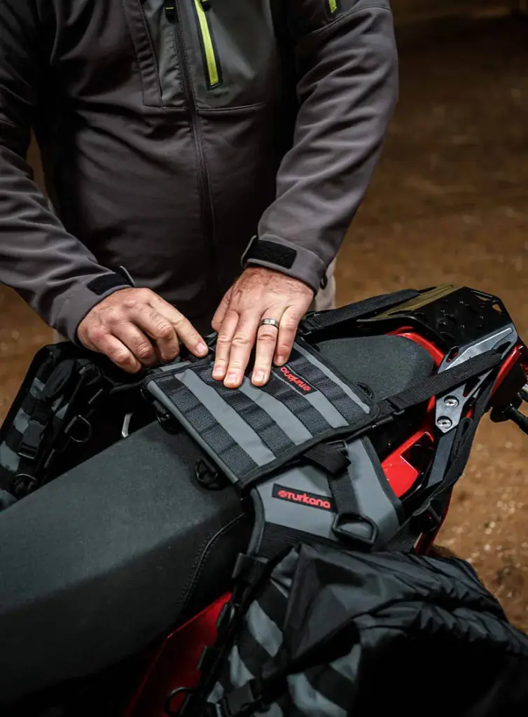 Hands strapping a Turkana Gear Madmule base onto a motorcycle seat